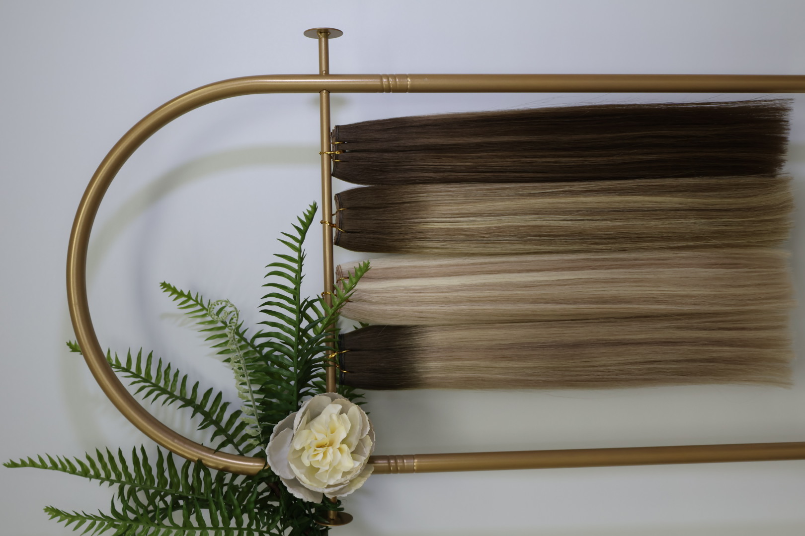 Brass display rack with curated balayage, blonde, and brunette extension swatches accented by ferns and a cream peony